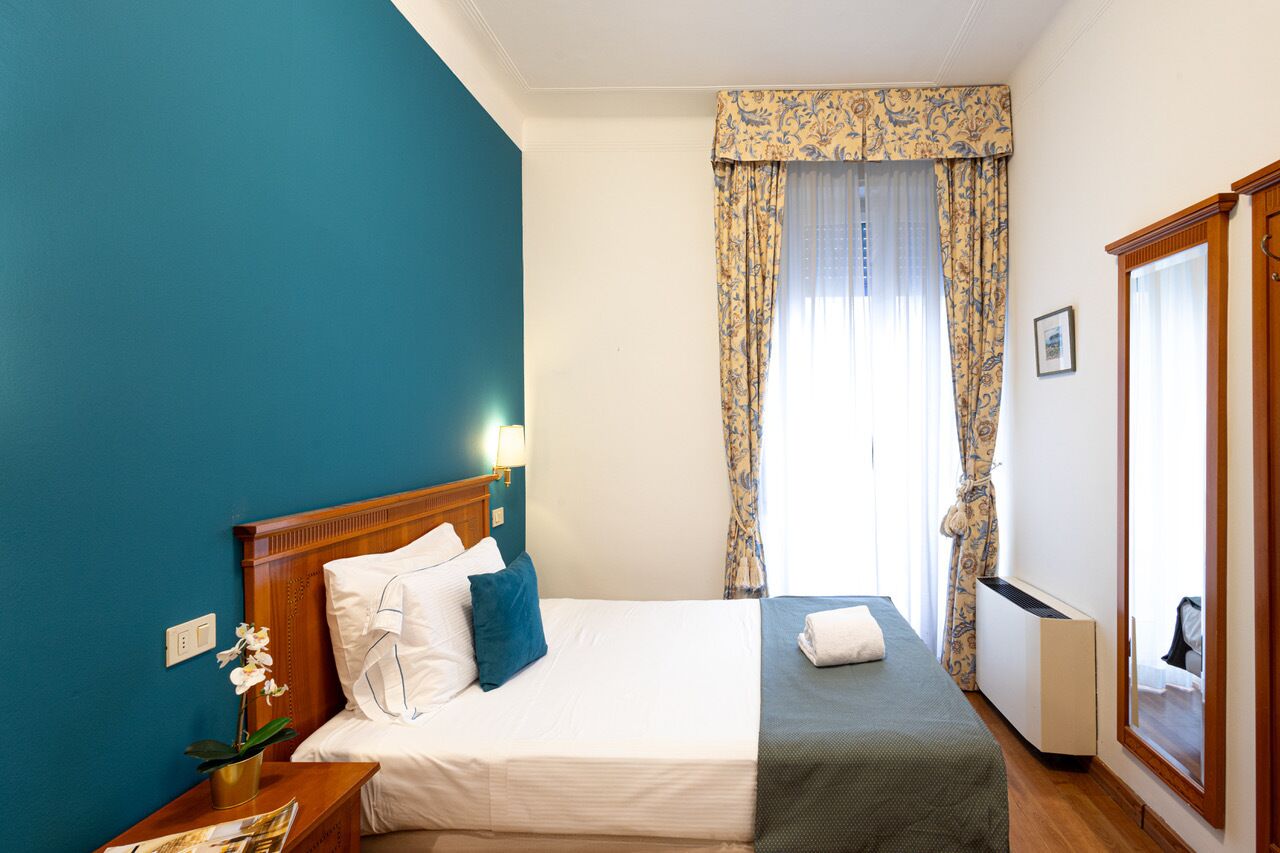 A hotel room with a bed, a nightstand with a lamp, a radiator, and a window with curtains.