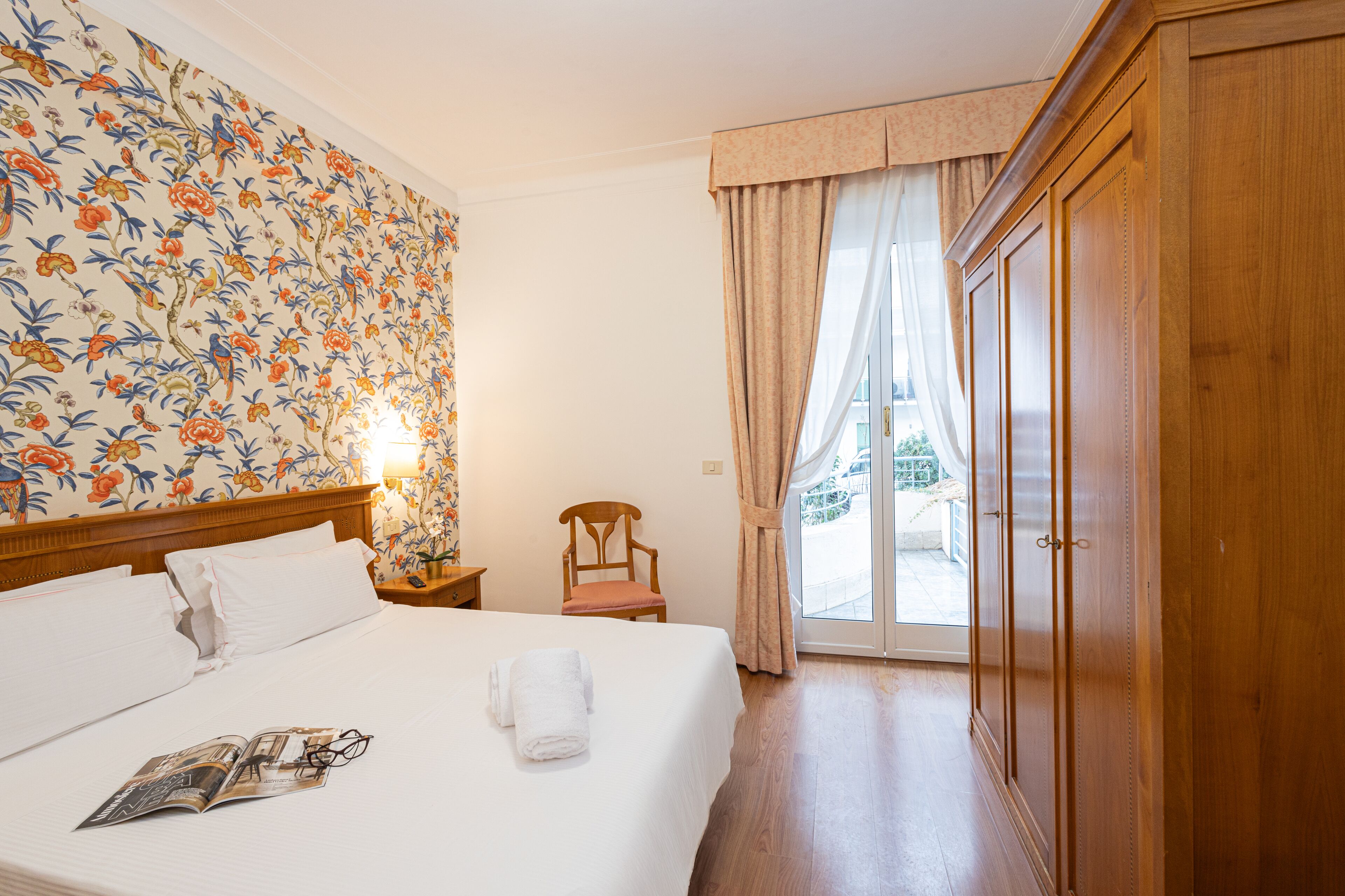 Habitación de hotel con cama, armario de madera, una silla y vistas a través de un ventanal.
