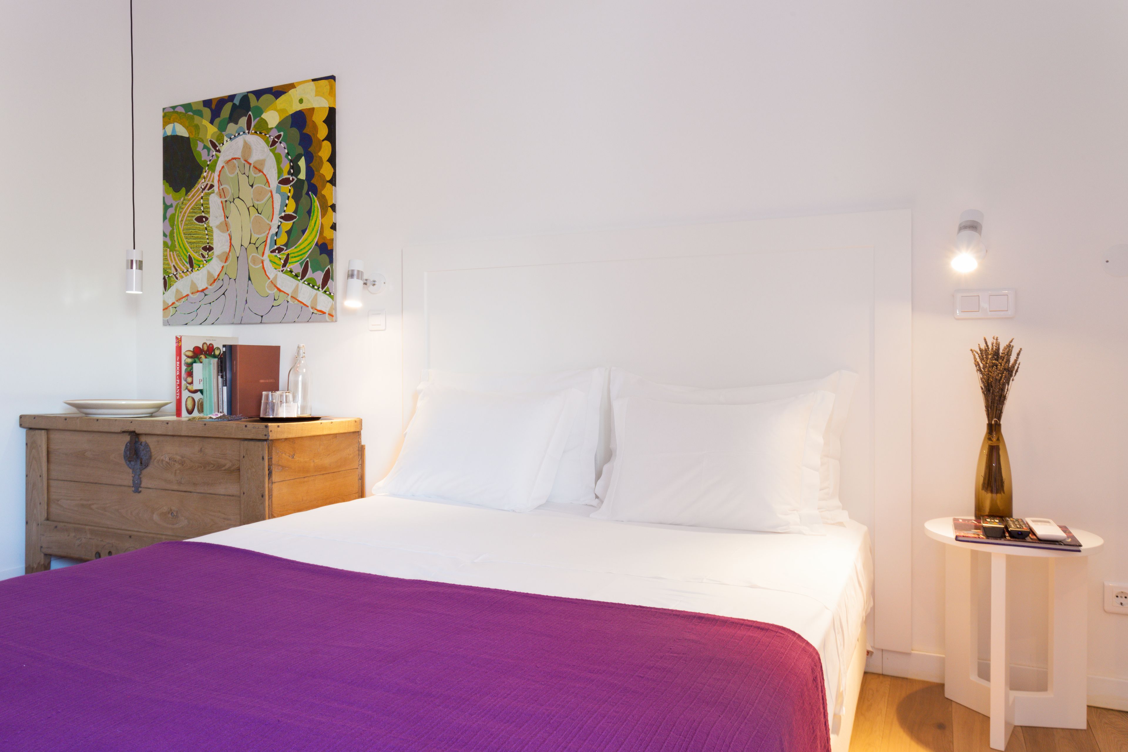 Un dormitorio con cama, cómoda, un cuadro, un florero y una mesita.