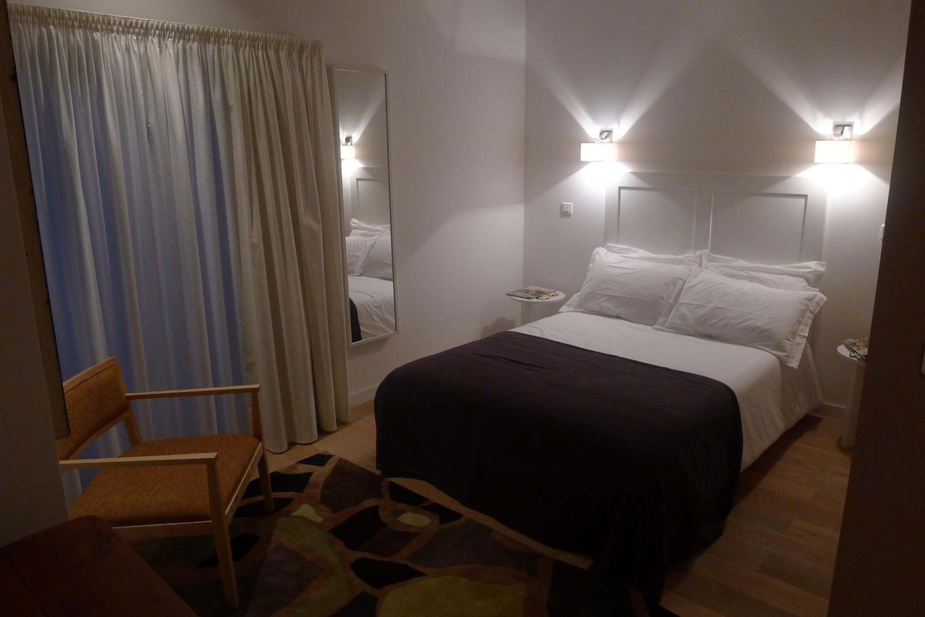A hotel room with a bed, a chair, a nightstand, and a mirror.