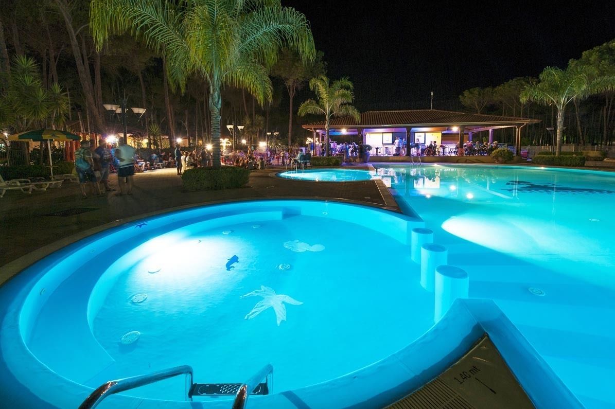 Un espace piscine nocturne comprenant plusieurs piscines, des palmiers et un espace de repos abrité.