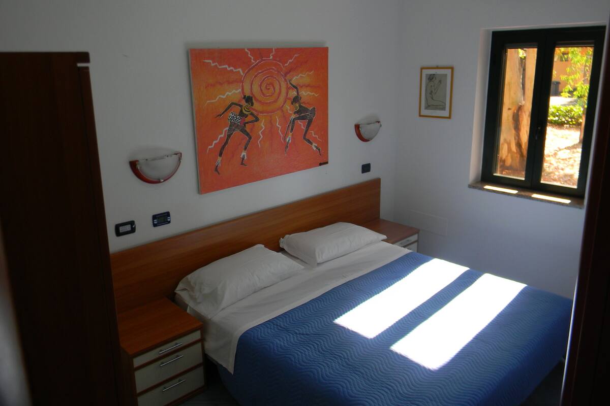 A bedroom with a bed, a nightstand, a wall-mounted artwork, a window, and a wall-mounted light fixture.
