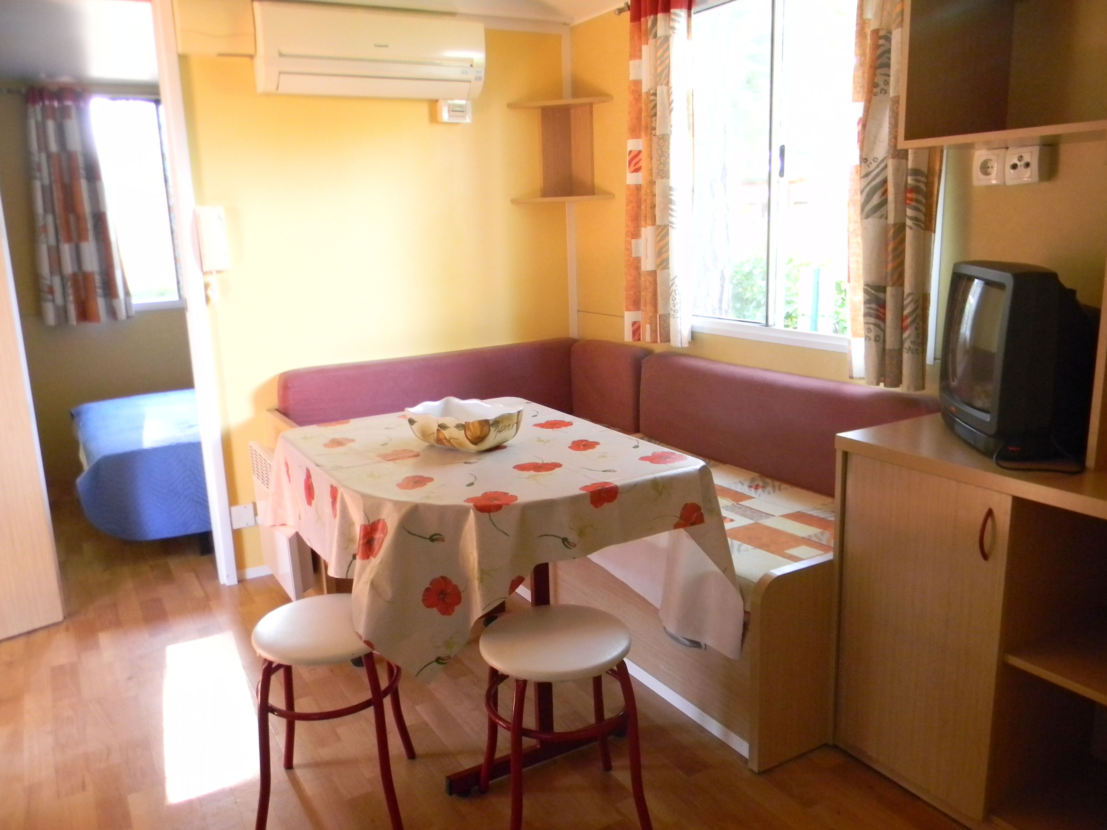 Un rincón comedor acogedor con una mesa cubierta por un mantel floral, dos taburetes, un banco acolchado y un televisor sobre un mueble de madera.