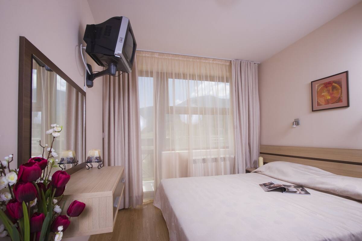 A hotel room with a bed, a bedside table, a mirror, a TV mounted on the wall, and a window with sheer curtains.
