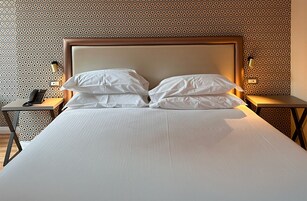 A neatly made bed with white linens and two pillows, a bedside table with a lamp, and a patterned wall in the background.
