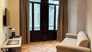 Ein modernes Hotelzimmer mit Holzboden im Fischgrätmuster, einer Couch, einem kleinen runden Tisch, einem Schreibtisch mit Lampe und einem Fernseher.