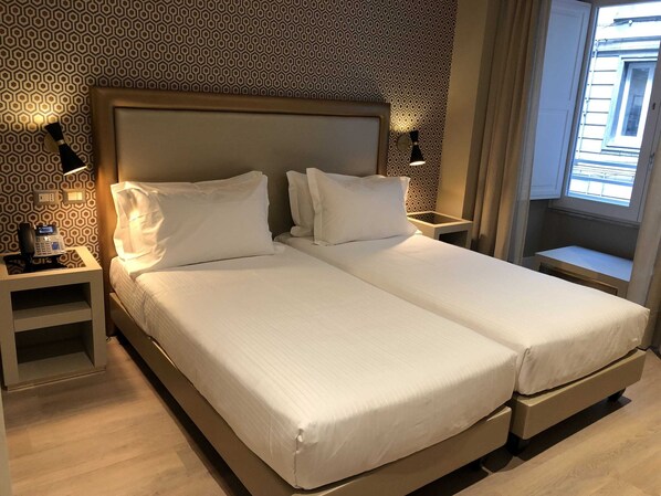 Una cama doble con sábanas blancas, una mesita de noche con lámpara y un papel tapiz estampado.