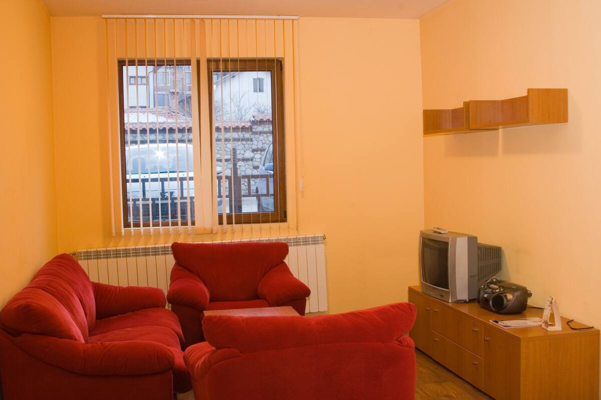 A living room with two red sofas, a wooden cabinet with a television and a speaker, and a window with blinds.