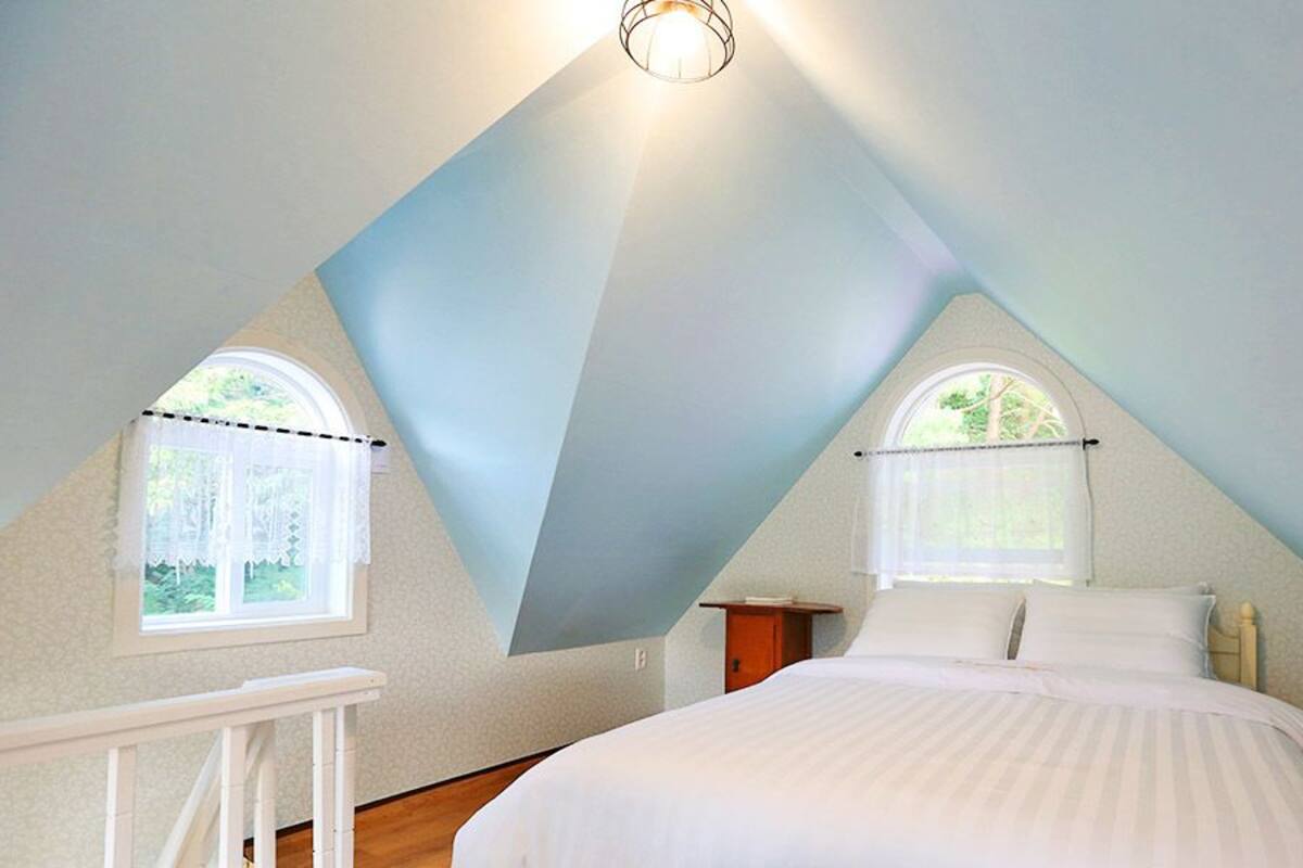 A bedroom with a sloped ceiling, two windows with curtains, a bed with white bedding, and a small bedside table.