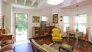 A living room with a television, yellow armchair, dining table, and chairs. There is a large window with blinds, a wooden cabinet, and a door leading outside. - Kismet Cottage (Pyeongchang)