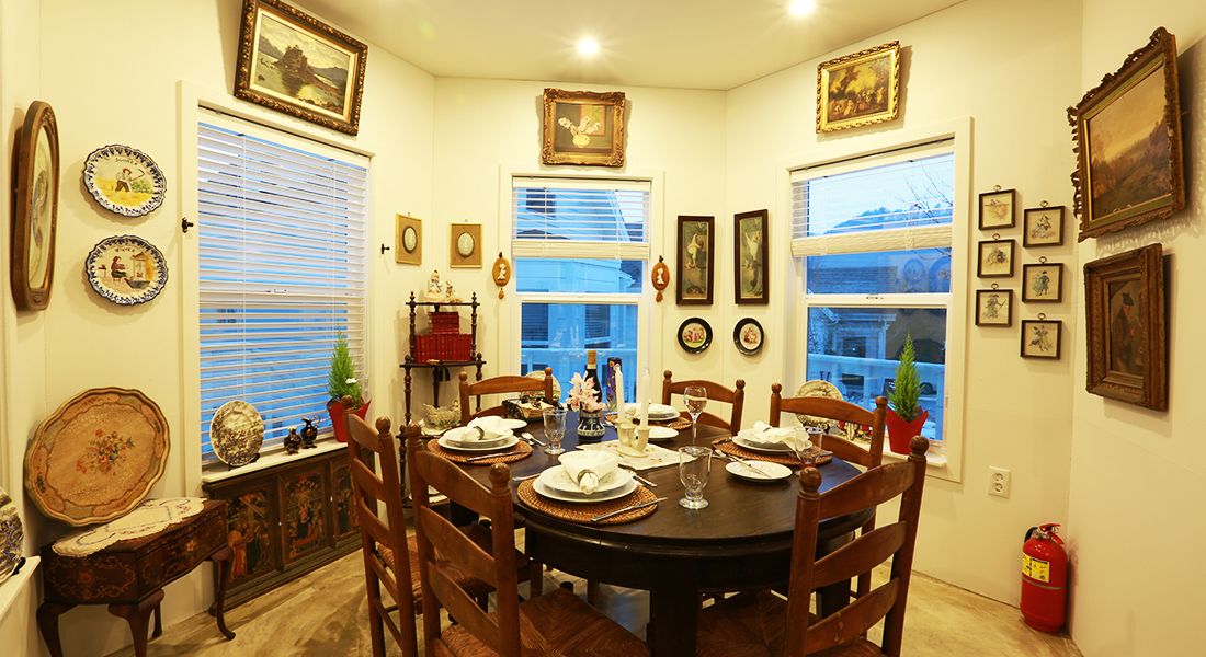 A dining room with a round table set for four, surrounded by wooden chairs. There are framed pictures on the walls, a window with blinds, and a fire extinguisher on the floor.