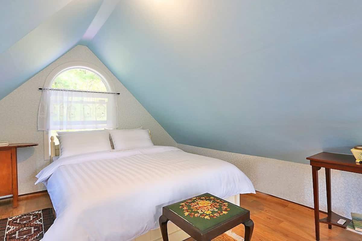 A bedroom with a bed, a window, a nightstand, a table, and a small stool.