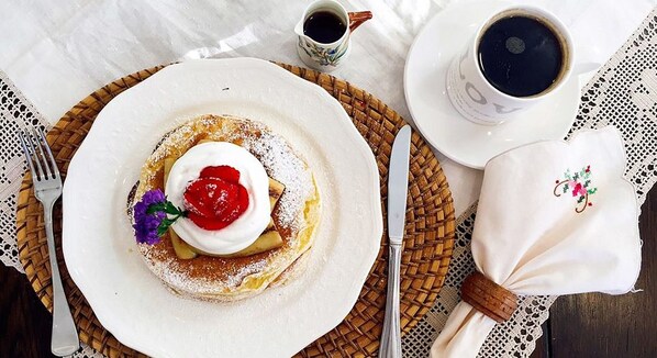 A breakfast setting with a plate of pancakes topped with whipped cream and a berry, a cup of coffee, and a napkin with a floral design. - Kismet Cottage (Pyeongchang)