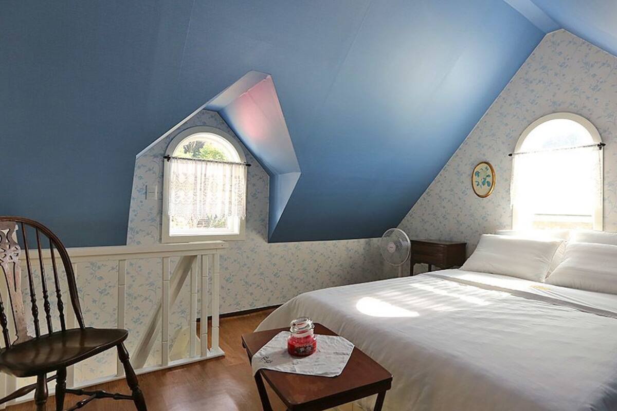 A bedroom with a sloped ceiling, a bed with white linens, a wooden chair, a small table with a jar, and a window with a curtain.