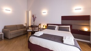 A hotel room with a bed, a sofa, a small table with a vase, and wall-mounted lights. - Savoy Riviera (Naples)