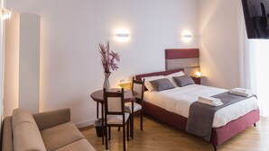 A modern hotel room with a wooden ceiling, a bed with a red headboard, a sofa, a small table with a vase, and a flat-screen TV mounted on the wall. - Savoy Riviera (Naples)