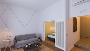 A modern living room with a grey sofa, a coffee table, and a floor lamp. There is a bed in the background and a radiator on the wall. - Savoy Riviera (Naples)