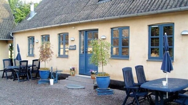 Un bâtiment à la façade beige, aux fenêtres bleues et au toit recouvert de tuiles. On y trouve des tables et des chaises à l’extérieur, ainsi que des plantes en pot.
