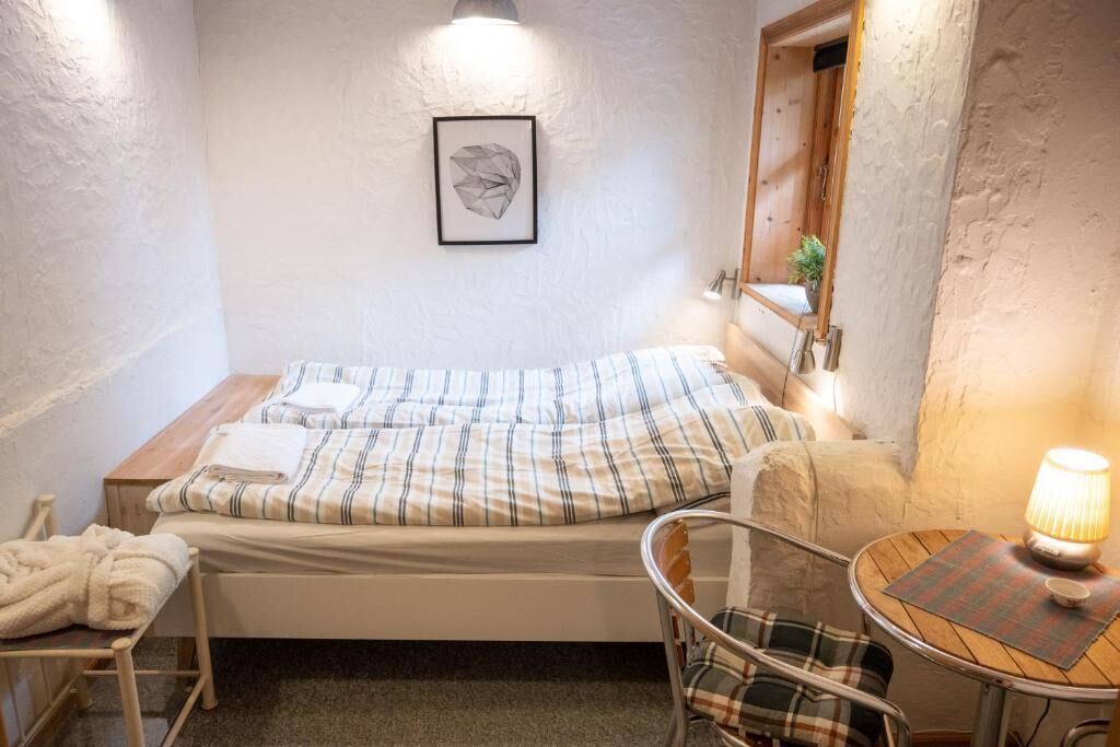 Una habitación pequeña y acogedora con una cama individual, una silla, una mesa con una lámpara y un cuadro en la pared.