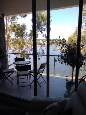 A balcony with a table and chairs, overlooking a body of water with trees and buildings in the background. - Marina View Apartment on the Maribyrnong River, Melbourne (Maribyrnong)