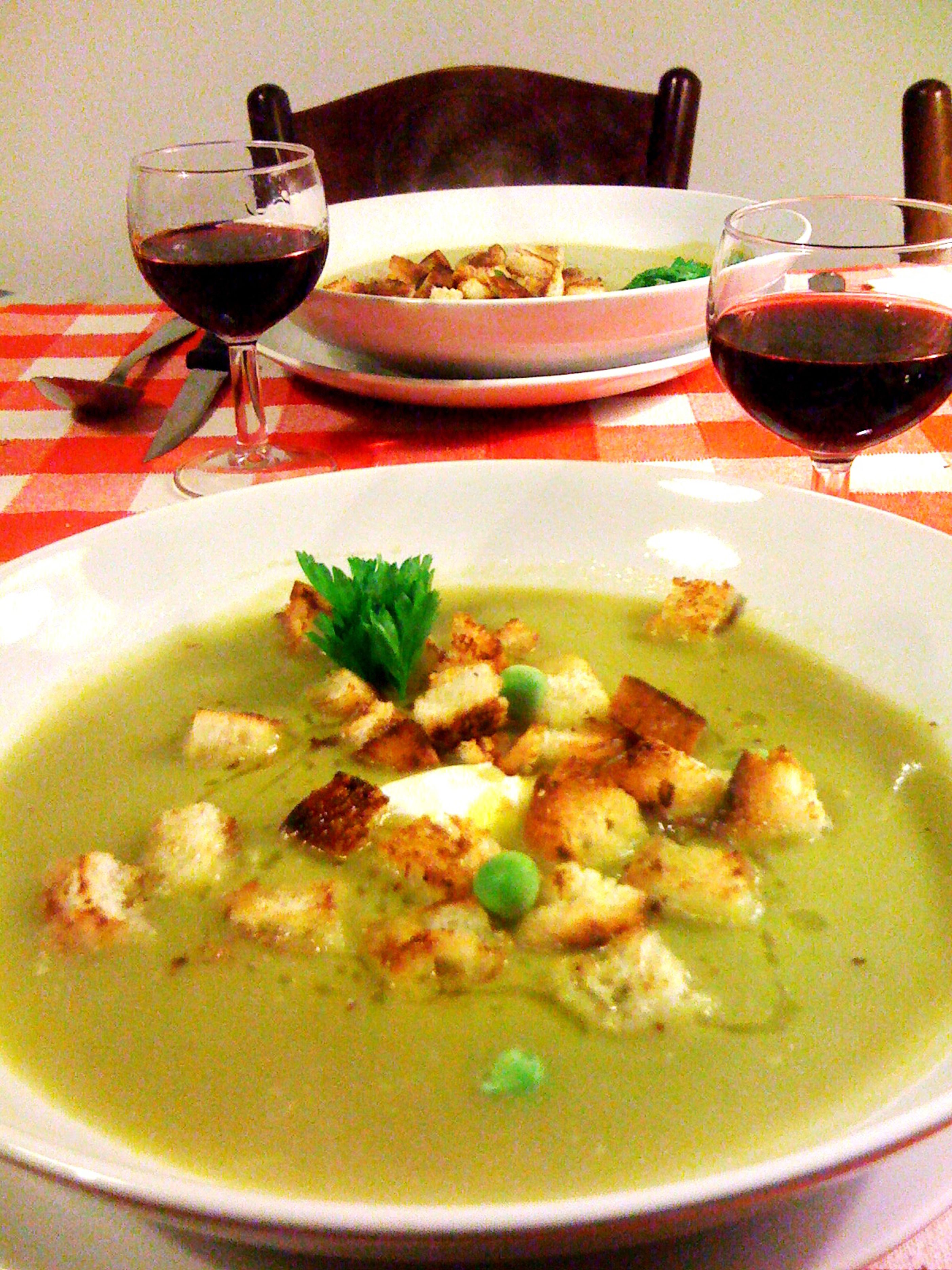 Un bol de soupe avec des croûtons et une garniture, deux verres de vin rouge et une nappe à carreaux.
