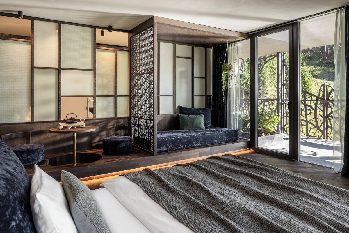 A modern bedroom with a large bed, a seating area with cushions, a small table with a teapot, and a view of greenery through large windows.