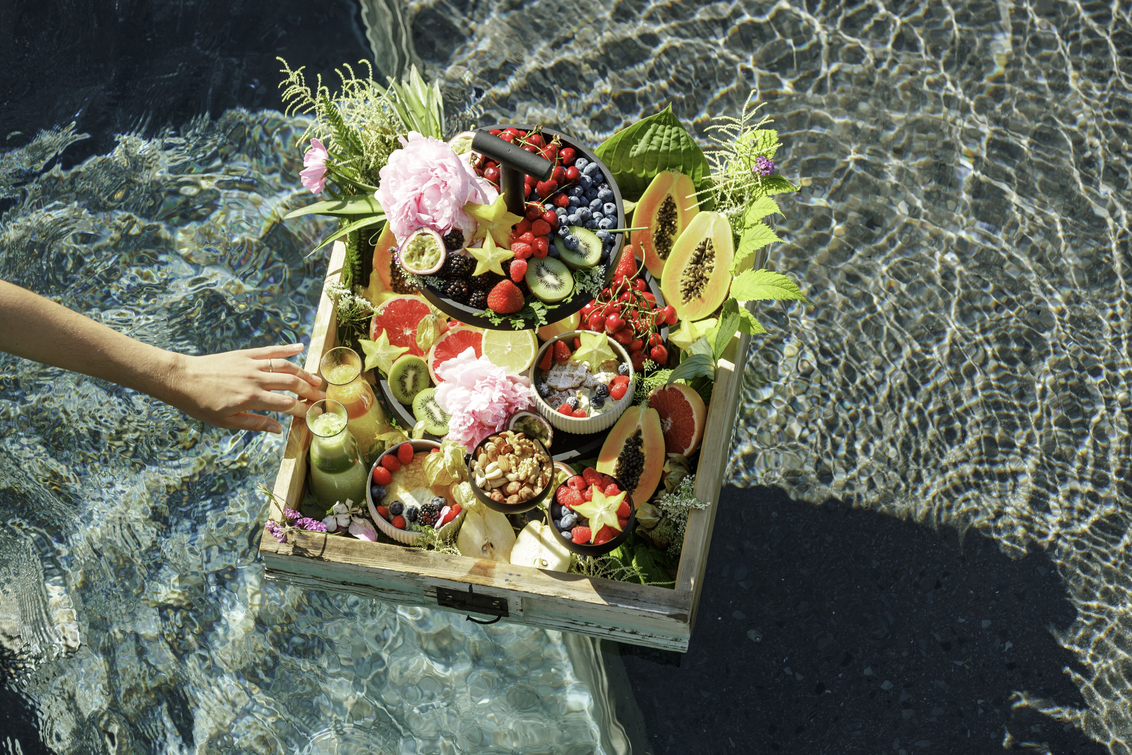 A wooden crate filled with fruits, vegetables, and flowers, held over a body of water.