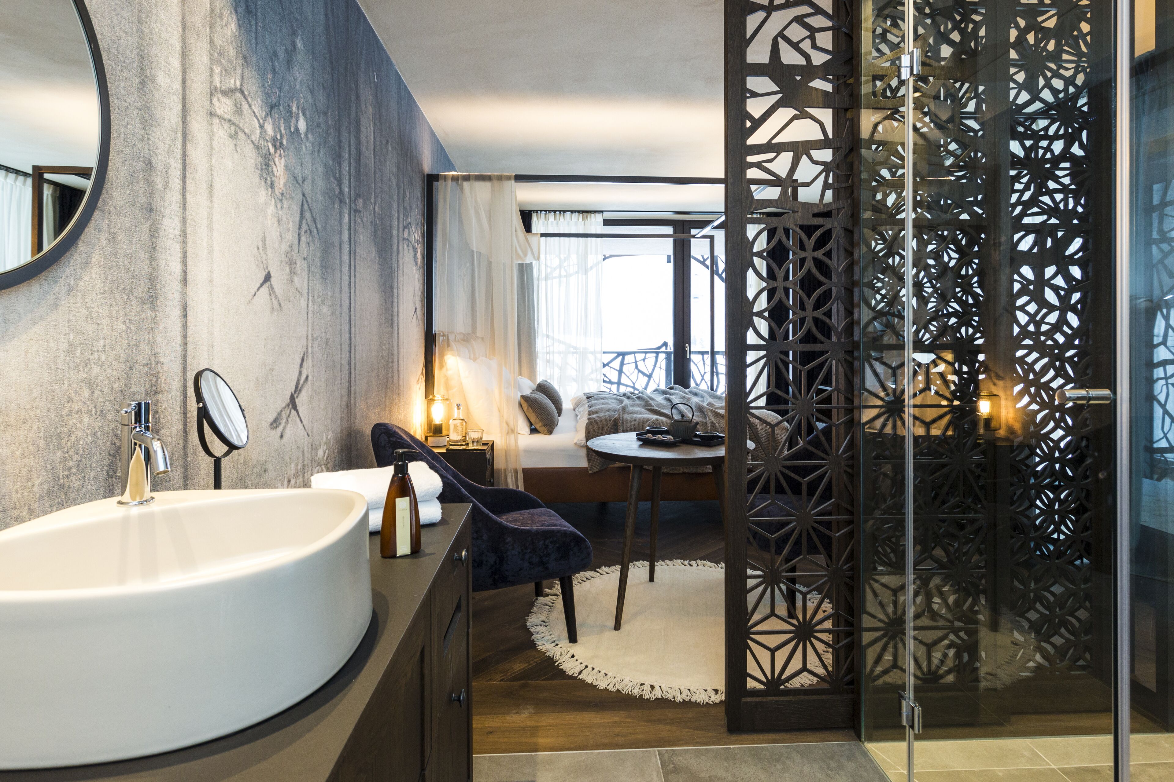 Ein modernes Badezimmer mit einem großen Waschbecken, einem Spiegel und einer Wandverkleidung. Ein Bett bietet Blick ins Freie, daneben ein kleiner Tisch mit Stuhl.