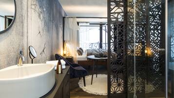 Ein modernes Badezimmer mit einem großen Waschbecken, einem Spiegel und einer Wandverkleidung. Ein Bett bietet Blick ins Freie, daneben ein kleiner Tisch mit Stuhl.
