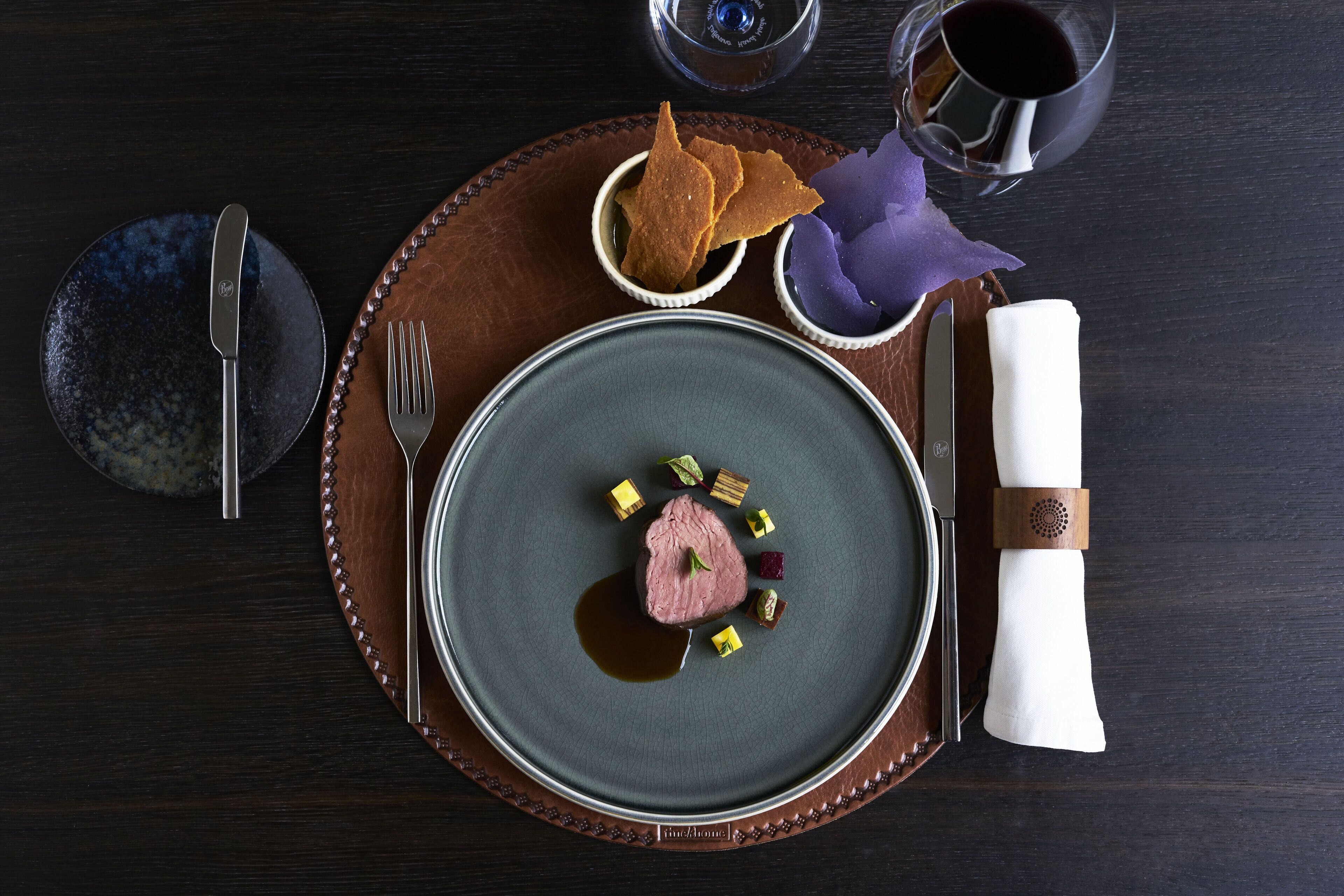 A formal dining setup with a plate of food, cutlery, and a glass of red wine on a dark wooden table.