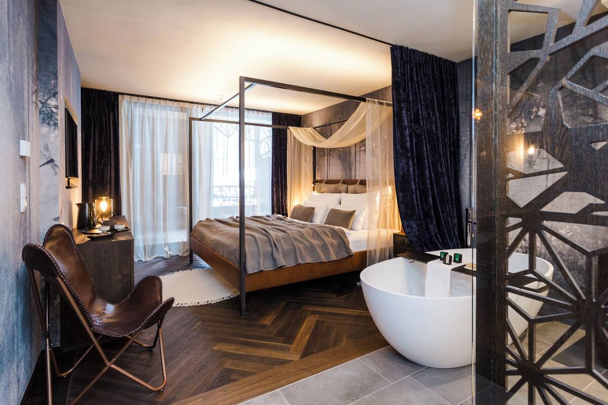 A modern bedroom with a four-poster bed, a large bathtub, and a leather chair.