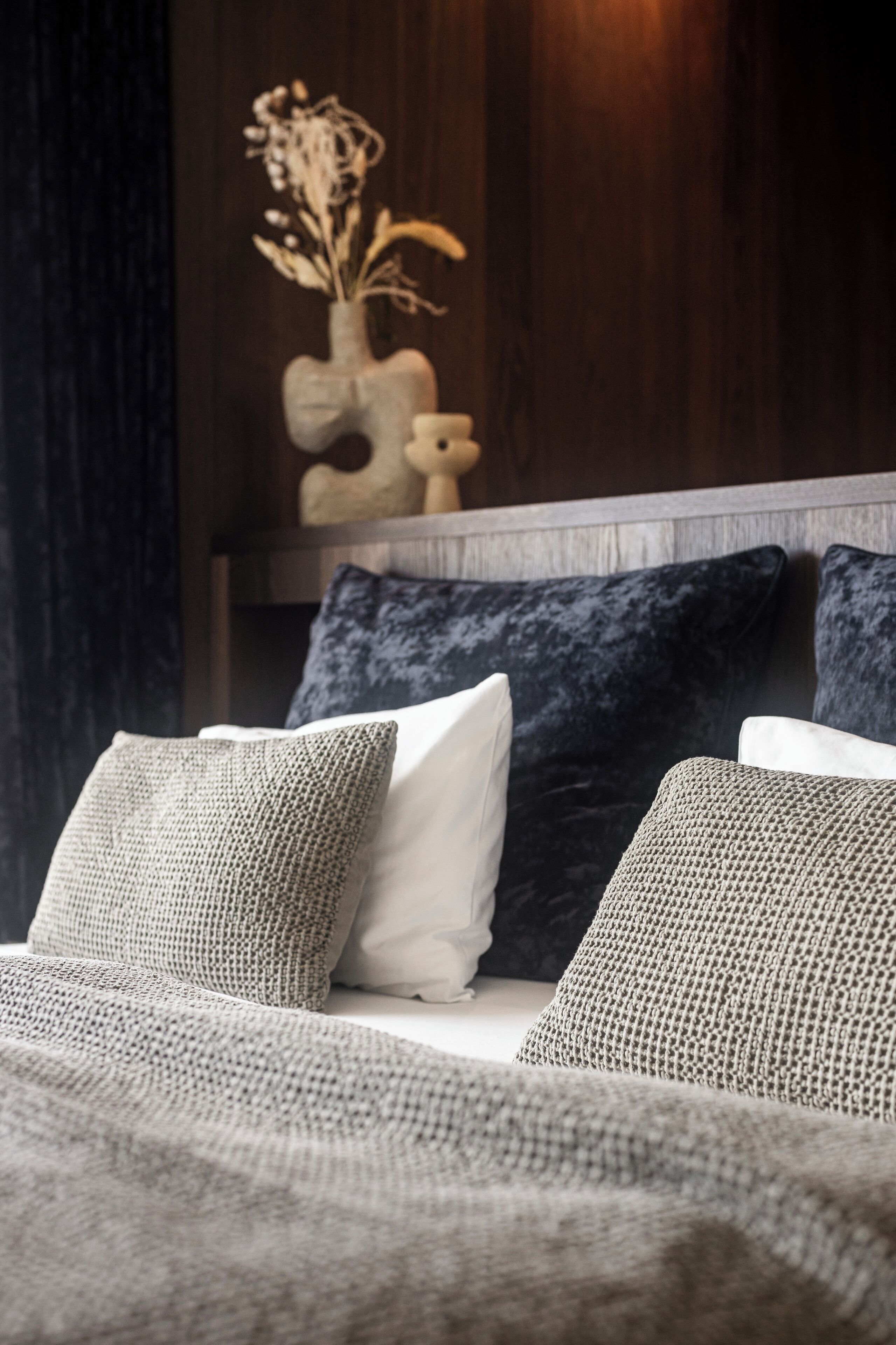 A bed with grey and white pillows, a dark headboard, and a decorative vase with dried flowers on a wooden shelf.