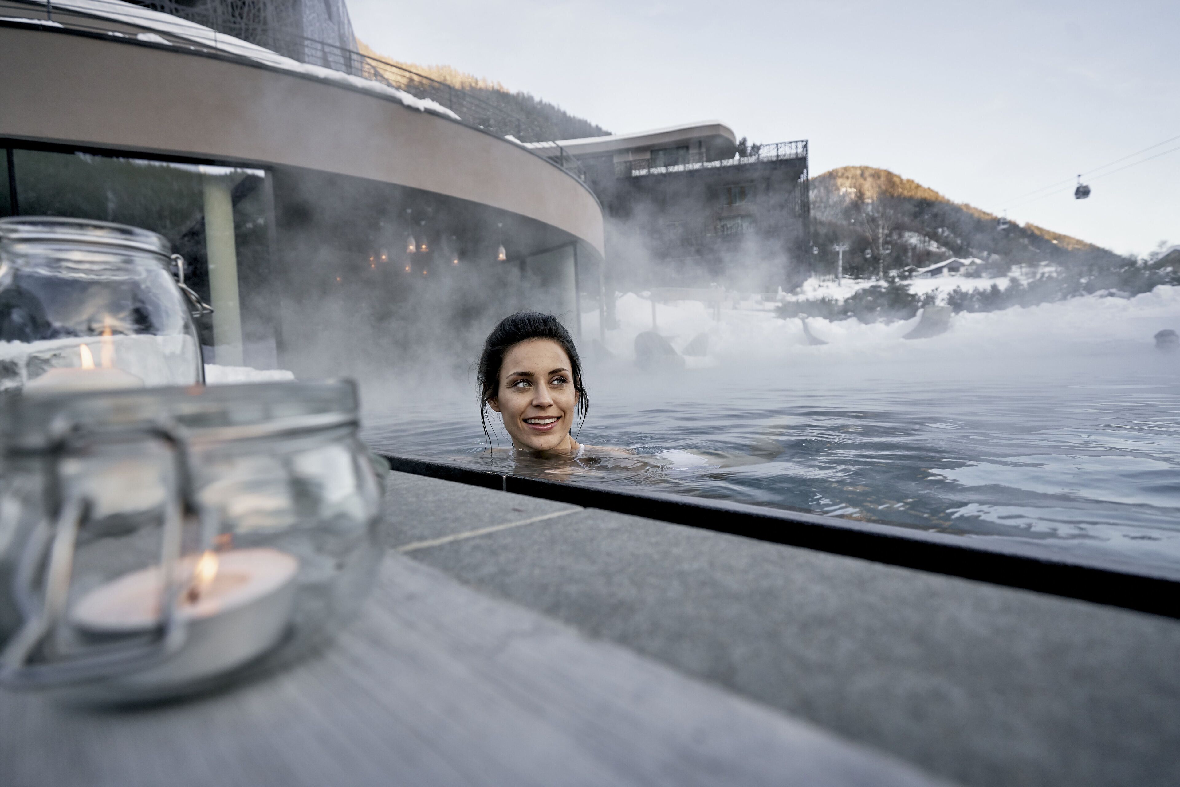 A person in a hot tub with steam rising, a glass jar with a candle, and a snowy mountain in the background.