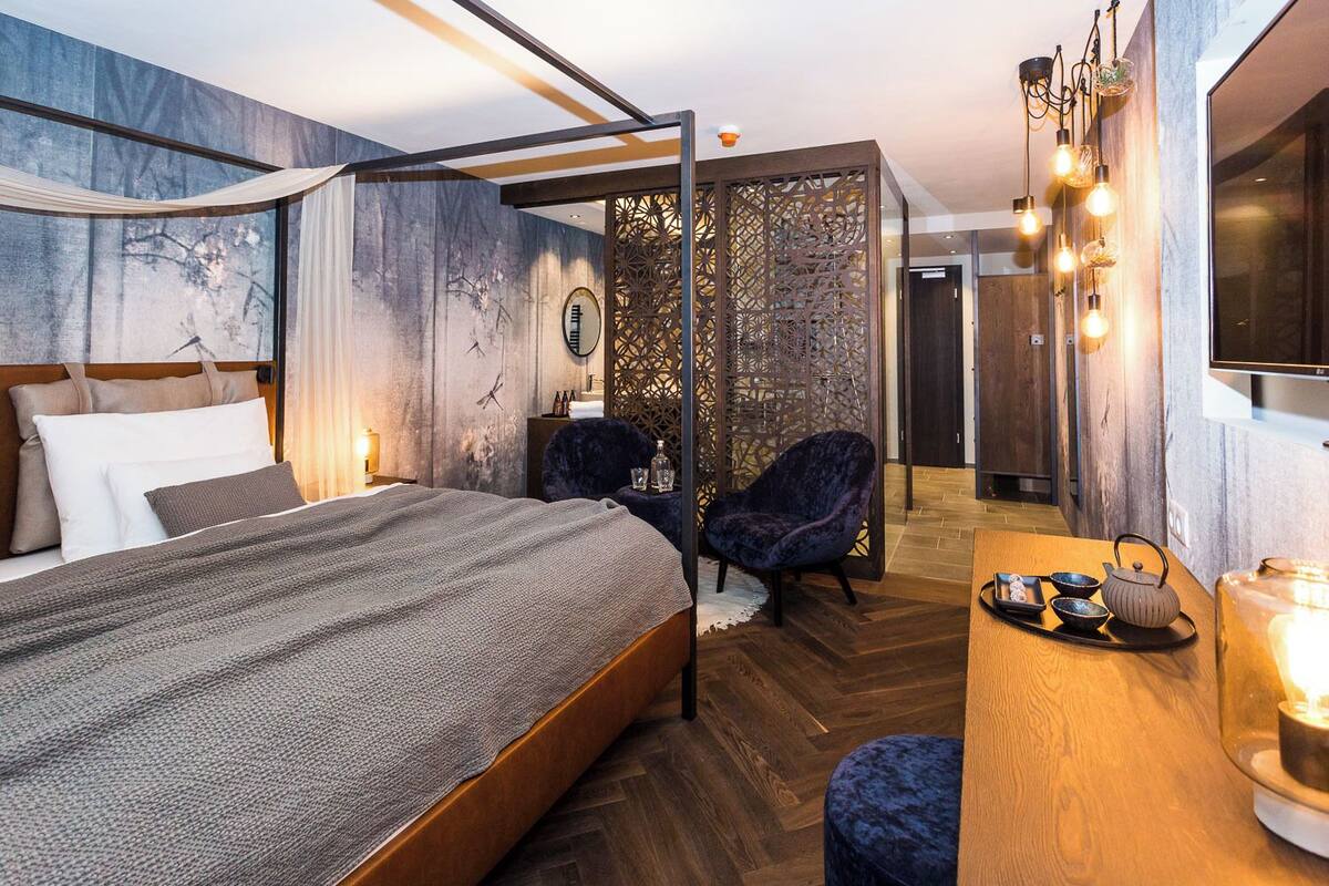 A modern hotel room with a large bed, two chairs, a desk with tea set, and a decorative screen.
