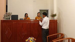A reception desk with a laptop, a name tag, and a plant. There are two chairs and a table with a bouquet of flowers. - Ocean Ngapali Lodge (Ngapali)