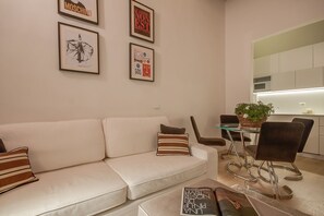 A modern living room with a white sofa, a glass coffee table, and a dining area with a glass table and metal chairs. There are framed artworks on the wall and a kitchen area with white cabinets and a microwave.