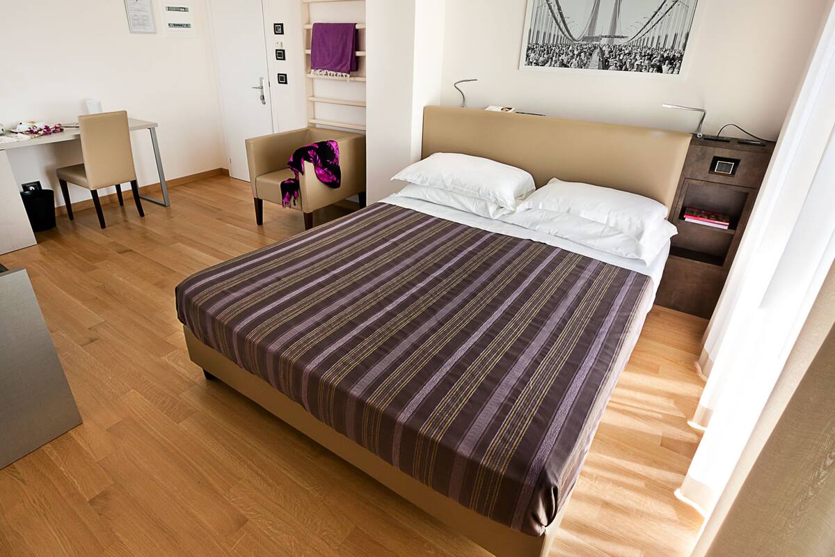 A hotel room with a large bed, a desk, a chair, a wardrobe, and a painting on the wall.