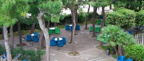 An outdoor seating area with blue and green chairs and tables, surrounded by trees and greenery.