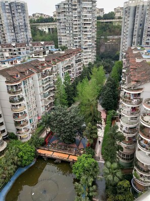 Un immeuble résidentiel de grande hauteur doté de nombreux balcons, d’une cour intérieure arborée et d’un petit pont enjambant un élément décoratif aquatique.