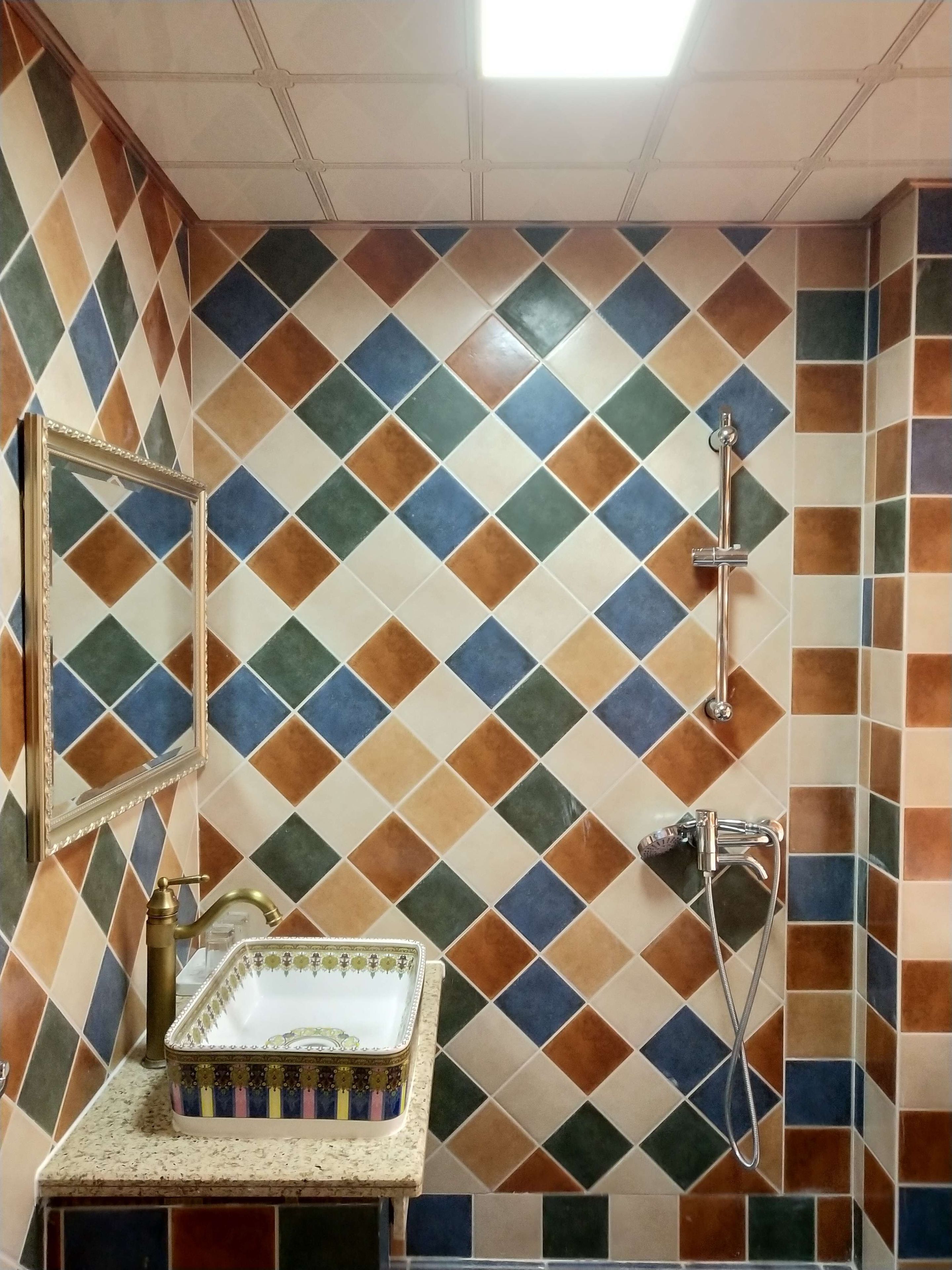 Un bany amb paret de rajoles amb dibuixos de rombes, un mirall, un lavabo amb un safata decorativa i una dutxa amb aixeta mànega.