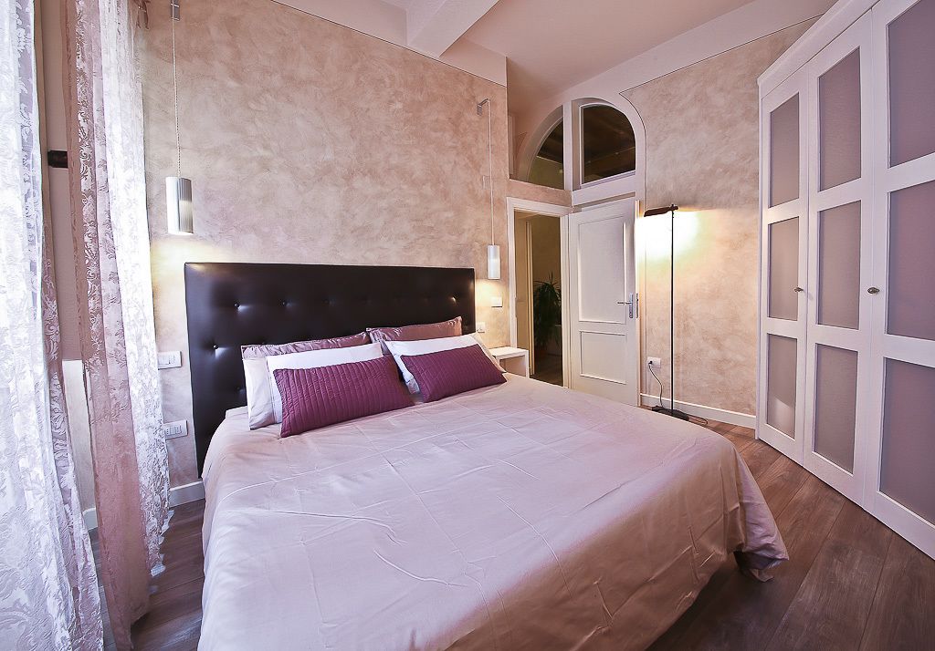 A bedroom with a large bed, a headboard, a wardrobe with glass doors, and a nightstand with a lamp.