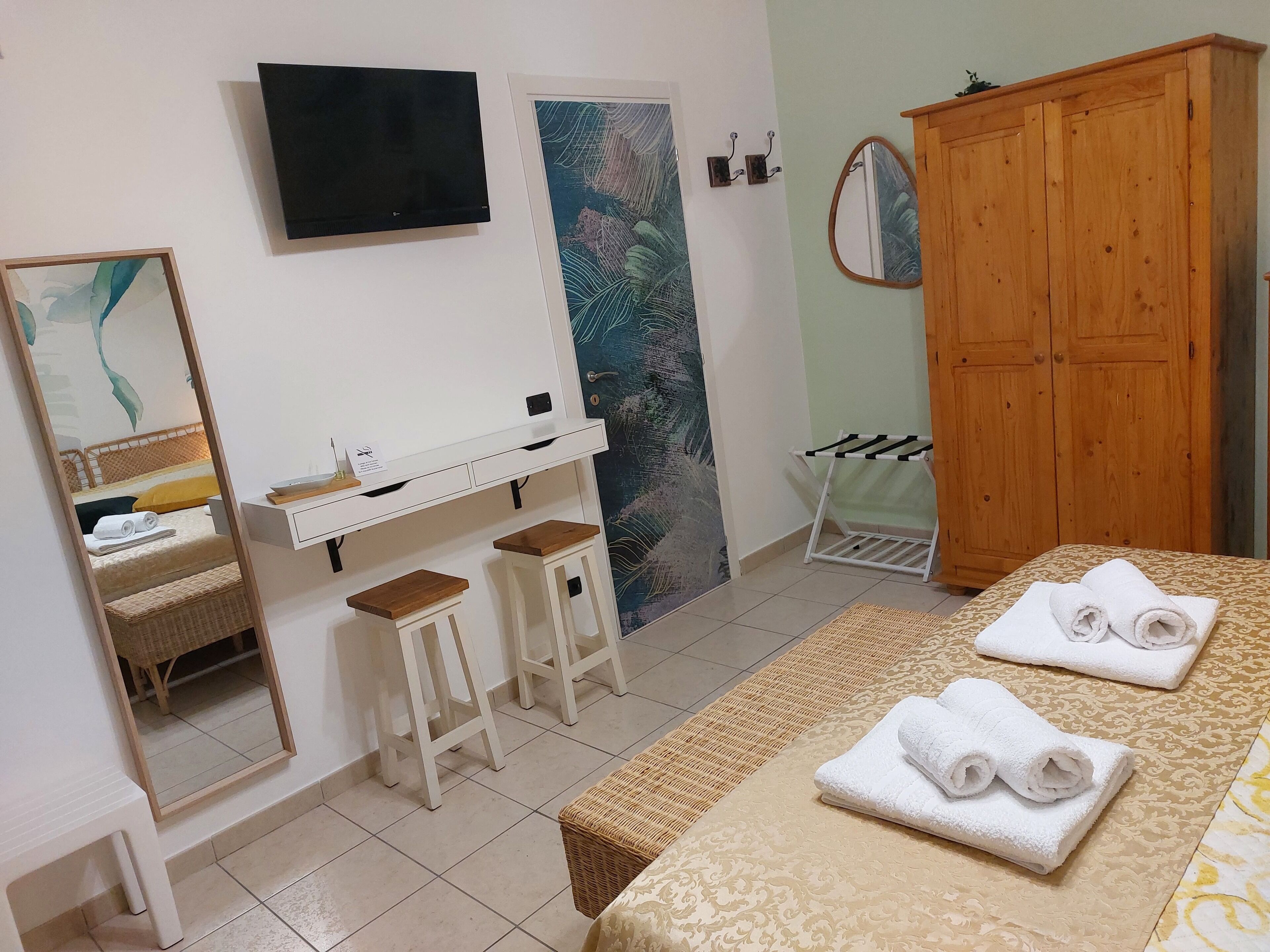 A hotel room with a bed, a wooden wardrobe, a TV, a painting, a mirror, and a small table with stools.