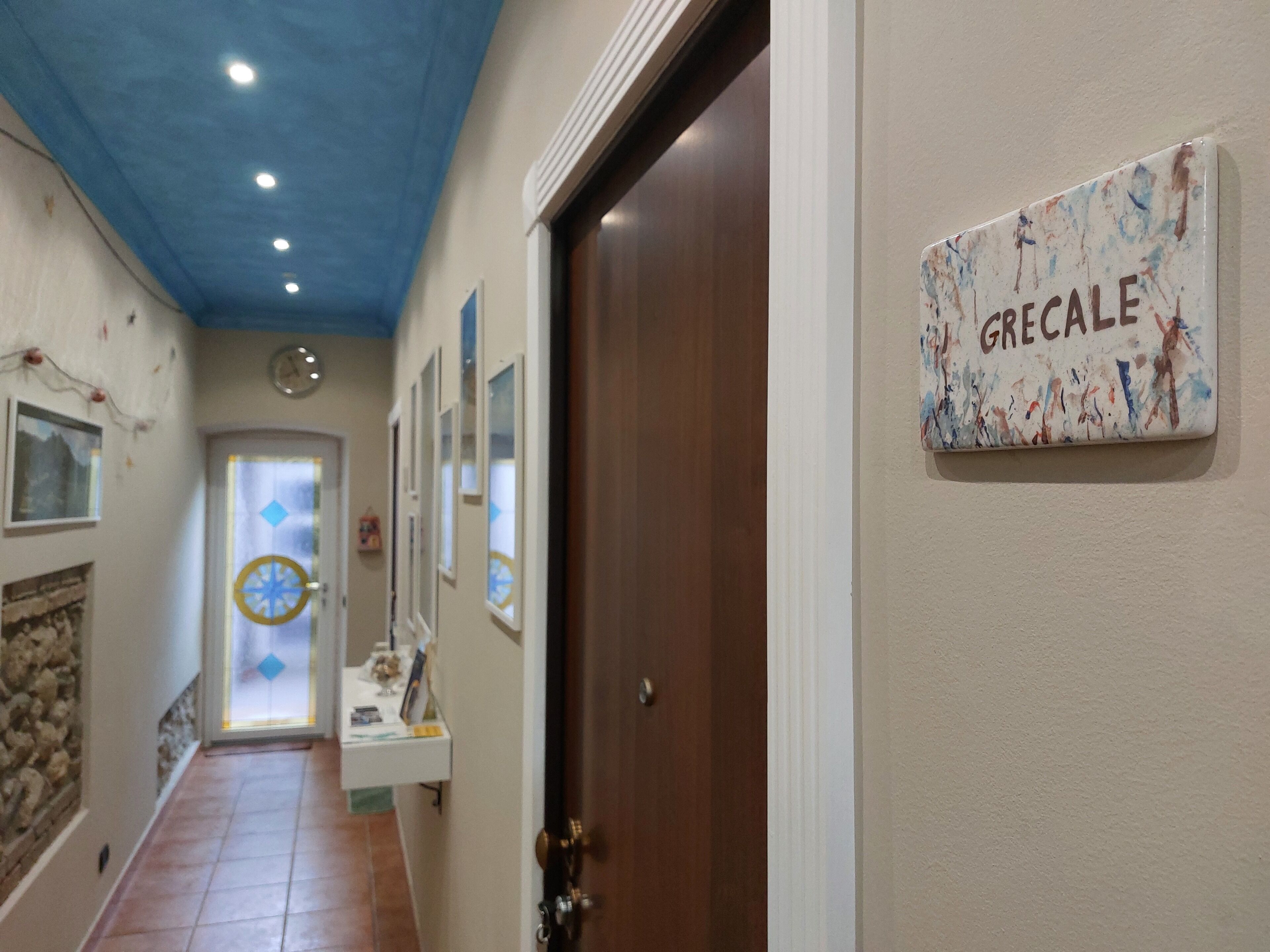 Un pasillo con una puerta identificada con la etiqueta 'GRECALE', un reloj de pared y un adorno mural.