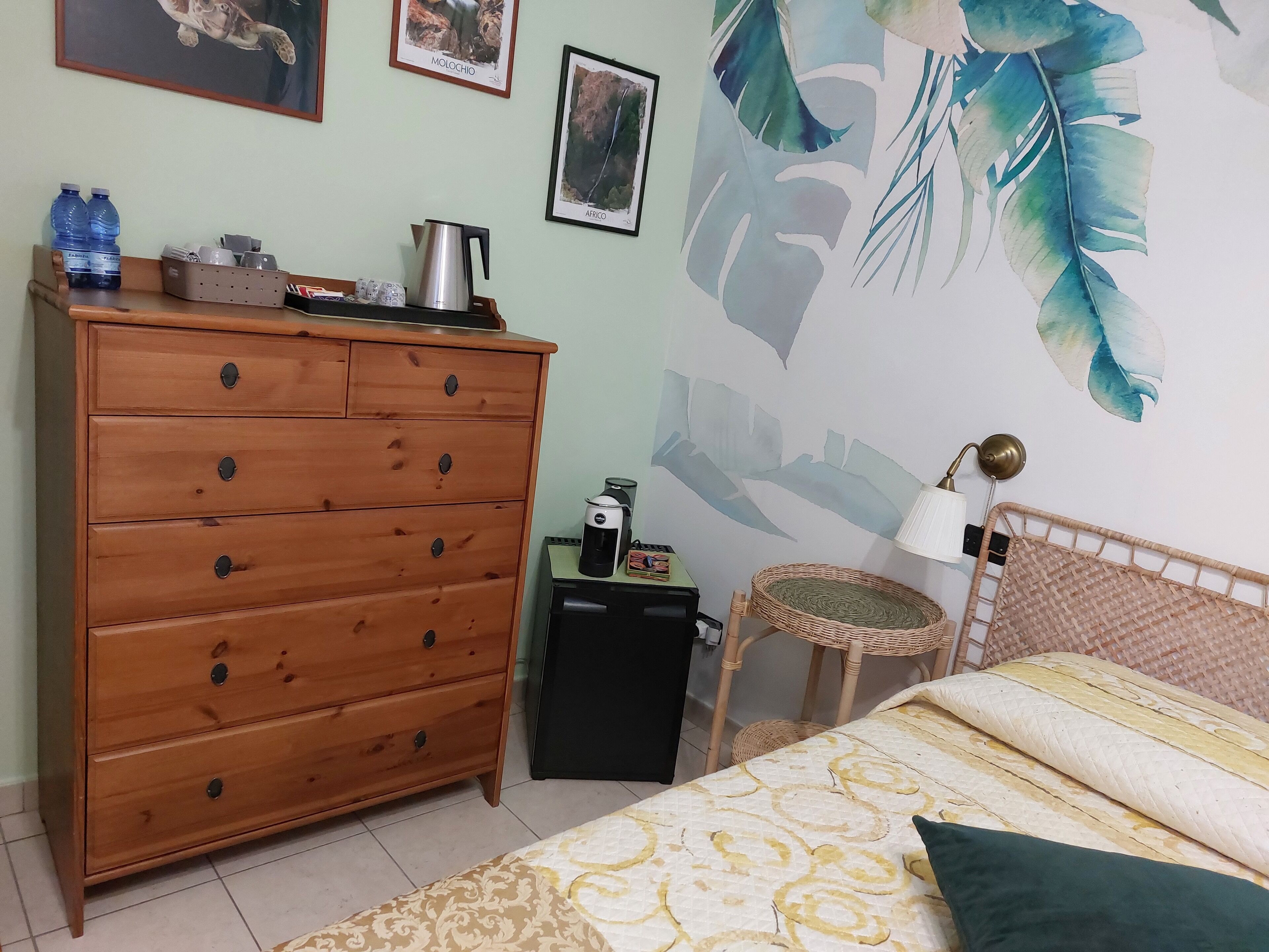 Un dormitorio con un aparador de madera, una cama con ropa de cama estampada, un cuadro de hojas tropicales en la pared y una mesita con un hervidor.