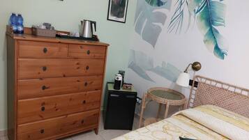Un dormitorio con un aparador de madera, una cama con ropa de cama estampada, un cuadro de hojas tropicales en la pared y una mesita con un hervidor.