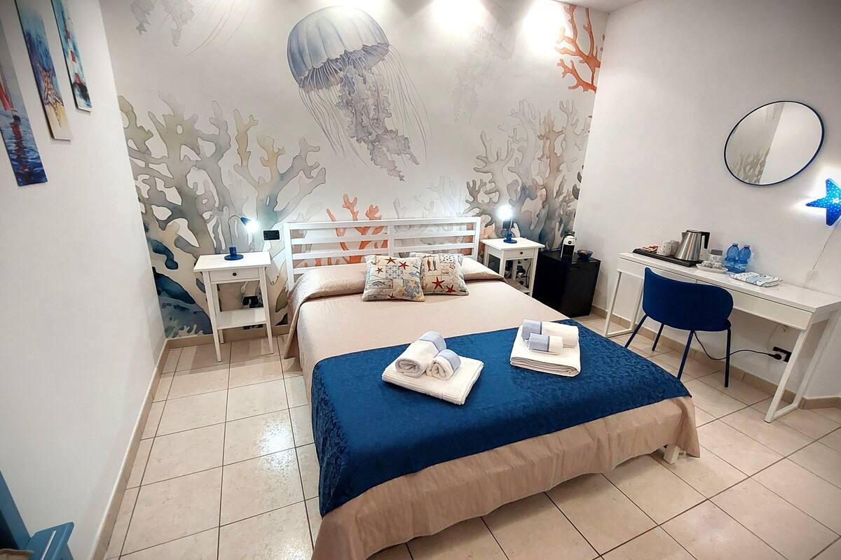 A bedroom with a bed, a desk, a chair, a nightstand, and a wall with a marine-themed mural.