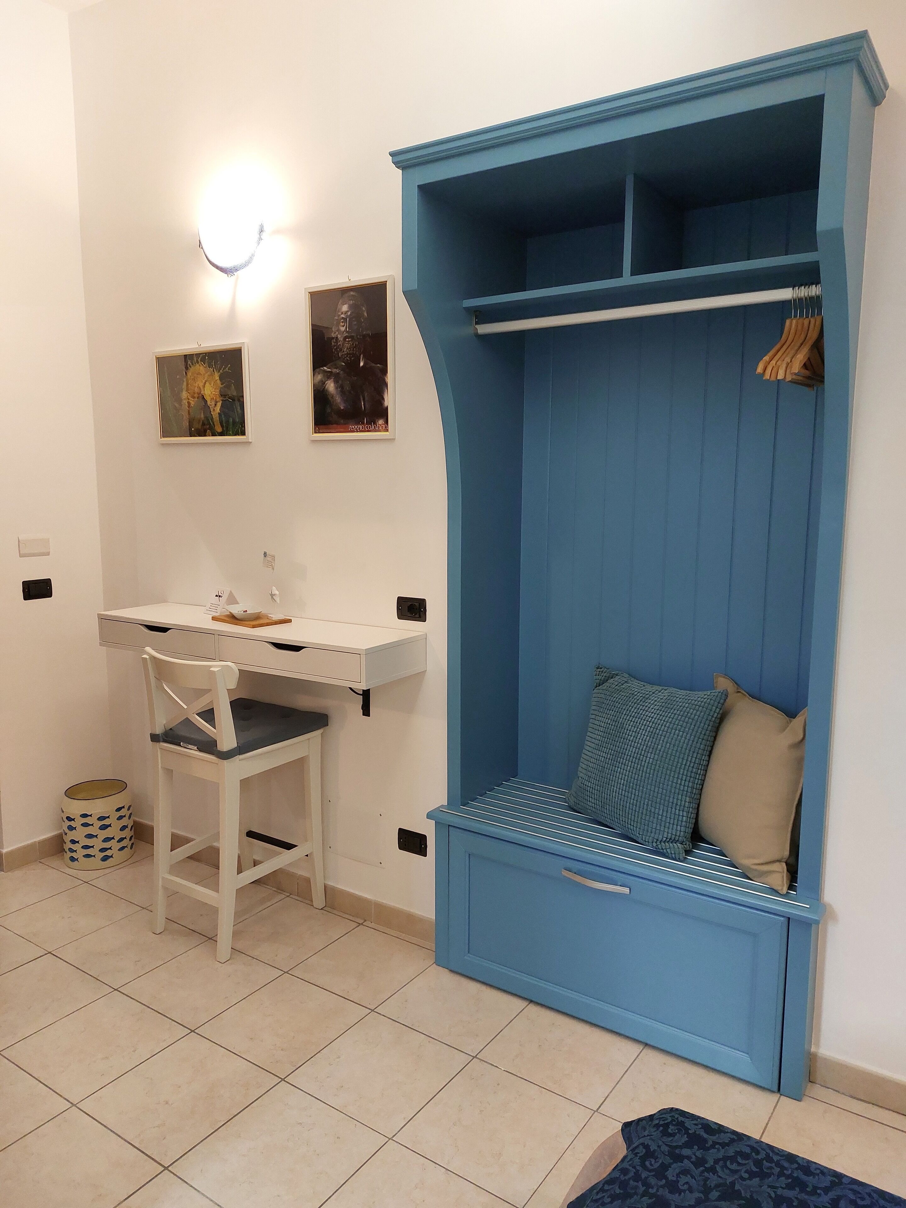 Une pièce de taille modeste, avec un bureau blanc, un banc bleu doté de rangements et une étagère fixée au mur.