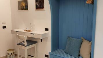 Une pièce de taille modeste, avec un bureau blanc, un banc bleu doté de rangements et une étagère fixée au mur.