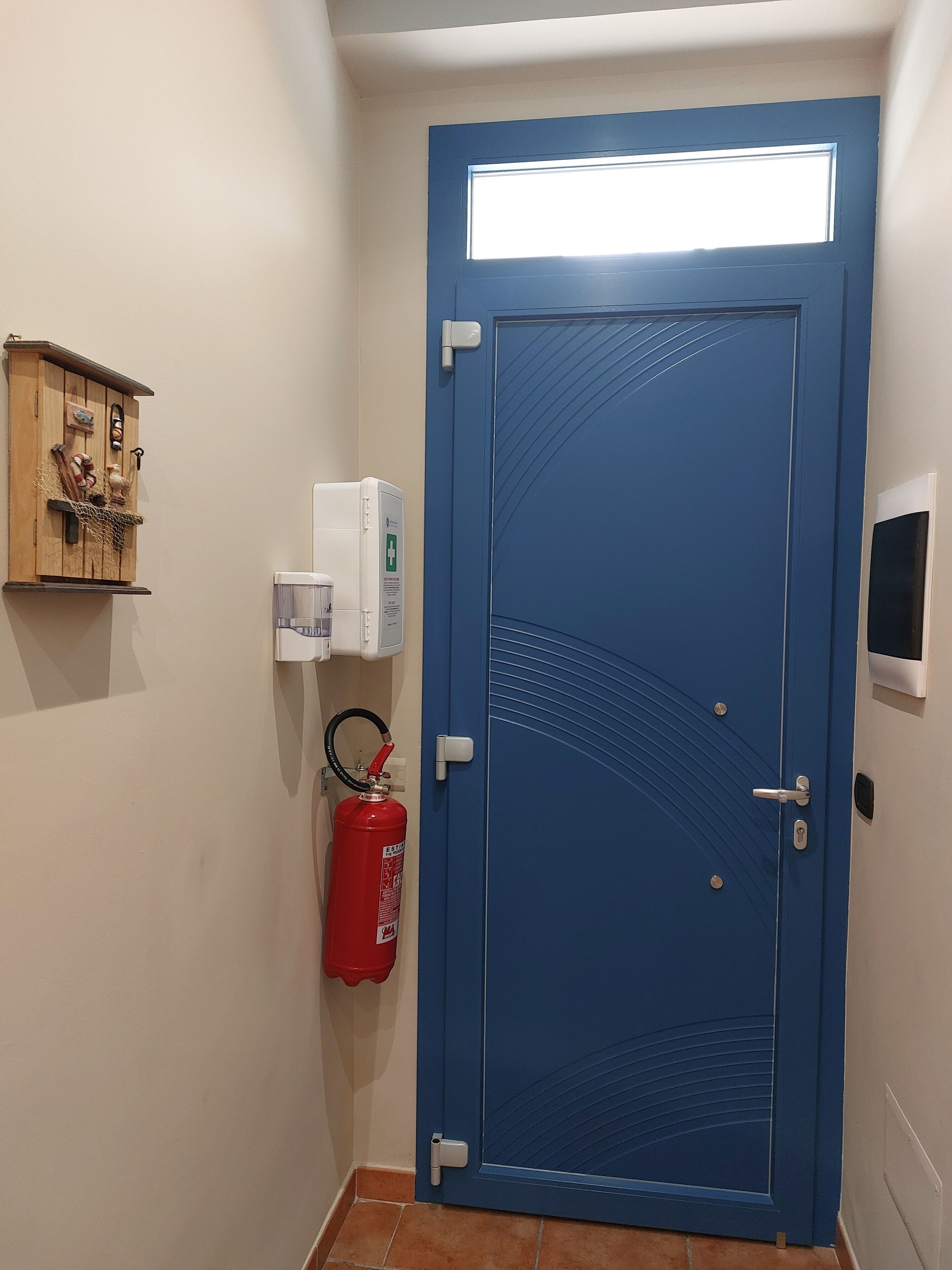 Une porte bleue avec une fenêtre, un extincteur, une petite étagère avec des objets décoratifs et un thermostat.