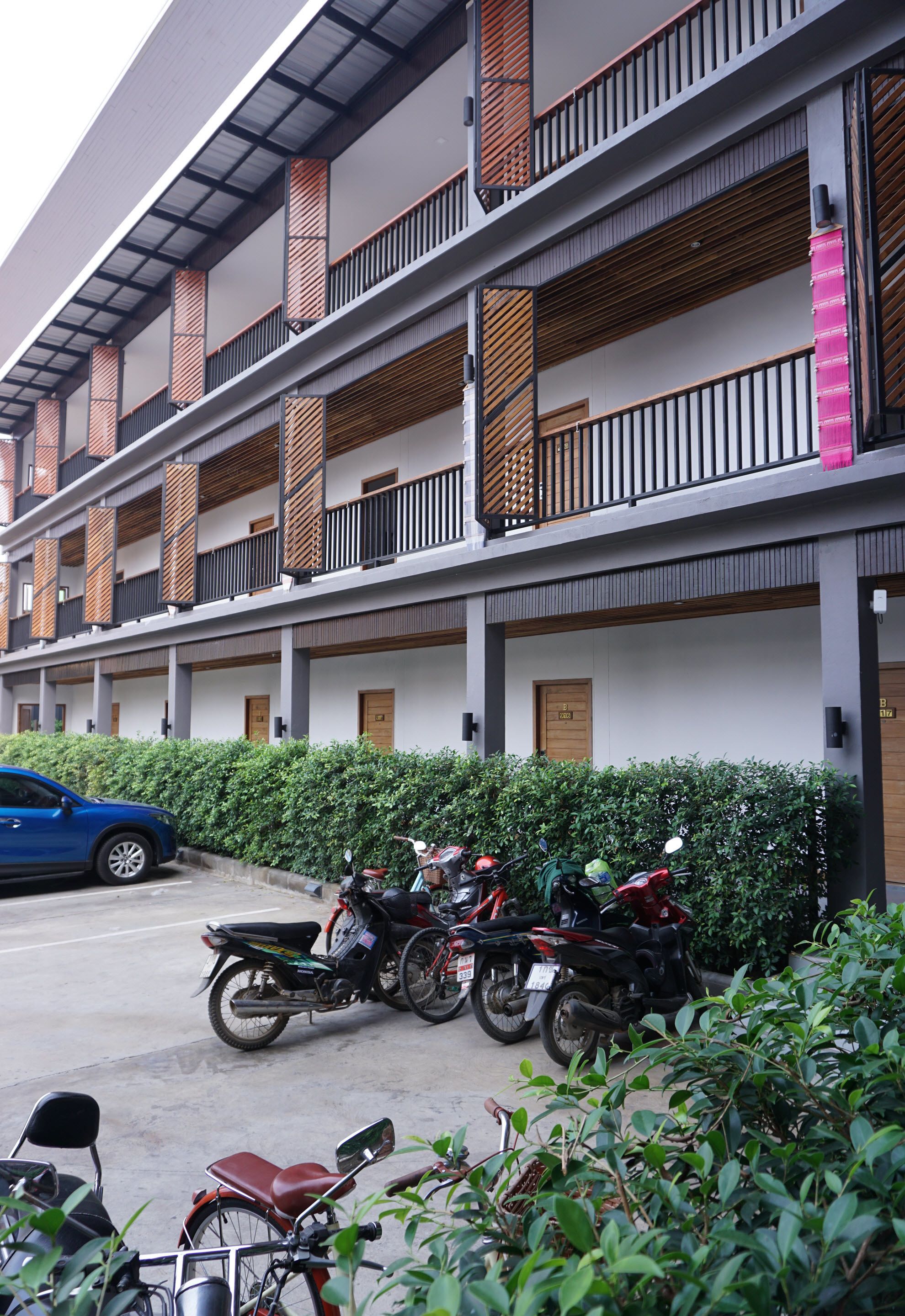 A multi-story building with balconies, a parking area with motorcycles, and a hedge in the foreground.